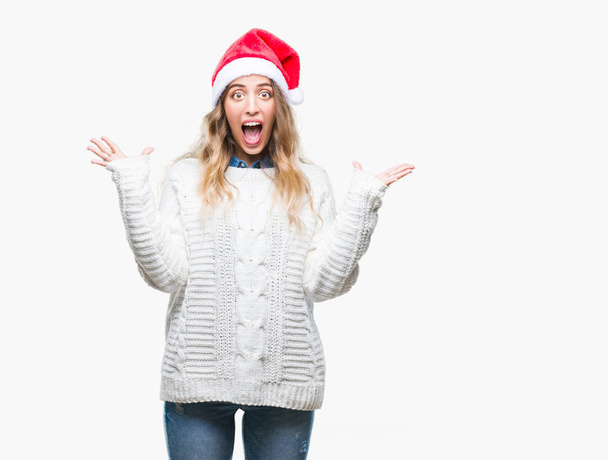 Piękna Młoda Blondynka kobieta kapeluszu Boże Narodzenie nad na białym tle obchodzi szalone i zdziwiony na sukces z podniesionymi rękami i Otwórz oczy krzyczy podekscytowany. Koncepcja zwycięzca - Zdjęcie, obraz
