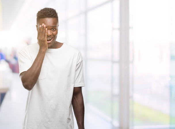 Jonge african american man die witte t-shirt die betrekking hebben op een oog met hand met vertrouwen glimlach op het gezicht en verrassing emotie. - Foto, afbeelding