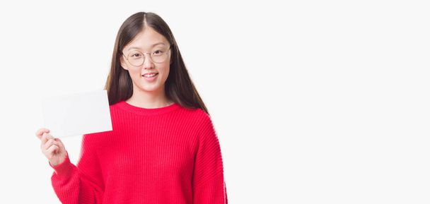 Joven mujer china sobre fondo aislado sosteniendo la tarjeta en blanco con una cara feliz de pie y sonriendo con una sonrisa confiada mostrando los dientes
 - Foto, imagen
