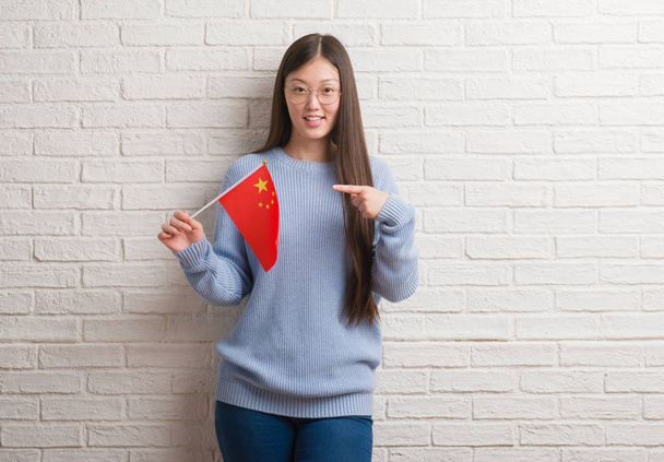 Fiatal kínai nő gazdaság zászlaja Kína nagyon boldog, rámutatva a kéz és az ujjak tégla falon - Fotó, kép