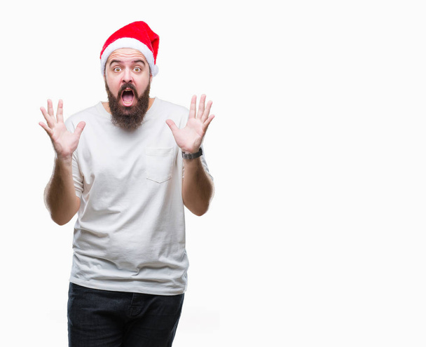 上クリスマスの帽子を着ている若い白人ヒップ男クレイジーを祝う背景を分離した腕を上げると成功のためびっくりし、叫んで興奮して目を開きます。勝者の概念 - 写真・画像