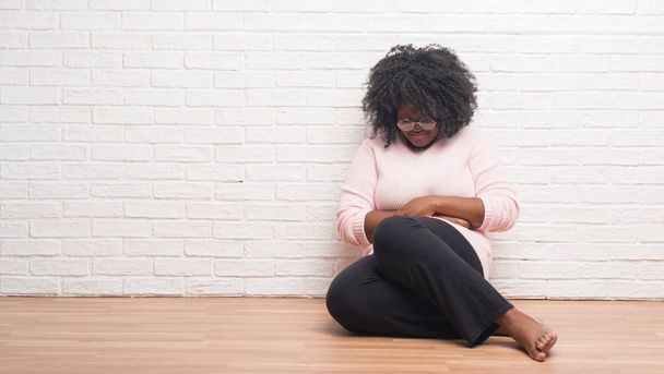 Jonge african american vrouw zittend op de vloer thuis met hand op de maag omdat indigestie, pijnlijke ziekte onwel. Ache concept. - Foto, afbeelding