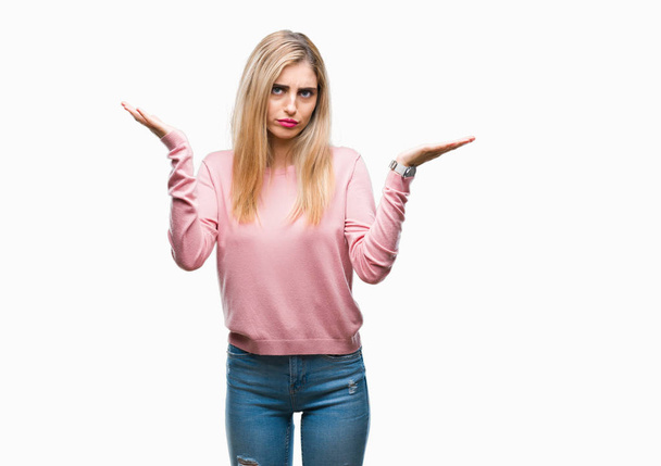 Young beautiful blonde woman wearing pink winter sweater over isolated background clueless and confused expression with arms and hands raised. Doubt concept. - Фото, изображение