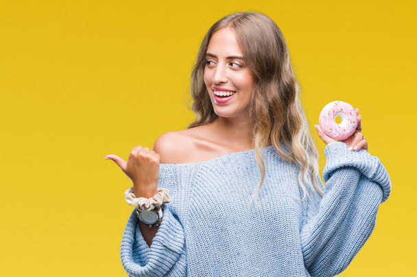 Beautiful young blonde woman eating pink donut over isolated background pointing and showing with thumb up to the side with happy face smiling - Foto, imagen