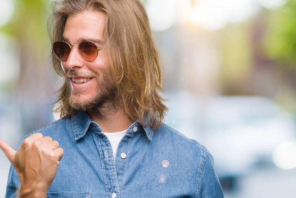 Young handsome man with long hair wearing sunglasses over isolated background smiling with happy face looking and pointing to the side with thumb up. - Photo, Image