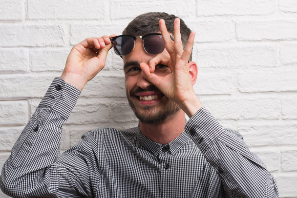 Hombre adulto joven con gafas de sol de pie sobre la pared de ladrillo blanco con la cara feliz sonriendo haciendo signo bien con la mano en el ojo mirando a través de los dedos
 - Foto, Imagen