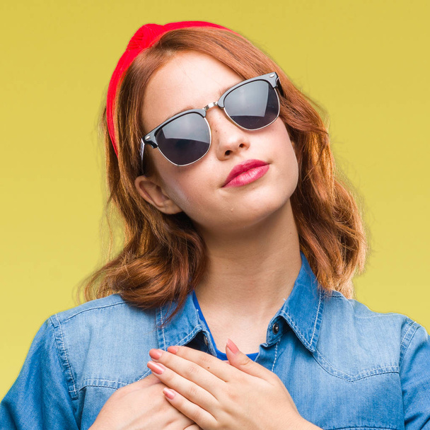 Young beautiful woman over isolated background wearing sunglasses smiling with hands on chest with closed eyes and grateful gesture on face. Health concept. - Фото, зображення