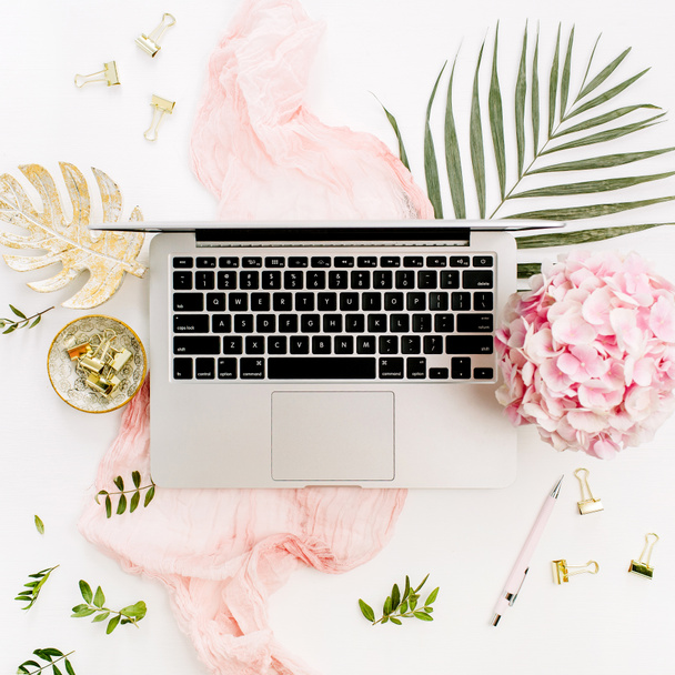 Сучасний домашній офісний стіл робочий простір з ноутбуком, букетом квітів рожевої гортензії, тропічним пальмовим листом, пастельно-рожевою ковдрою, листковою тарілкою та аксесуарами на білому тлі. Плоский прошарок, вид зверху
. - Фото, зображення