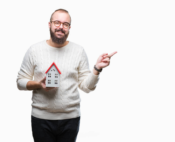 Young caucasian real state agent man holding house isolated background very happy pointing with hand and finger to the side - Foto, imagen