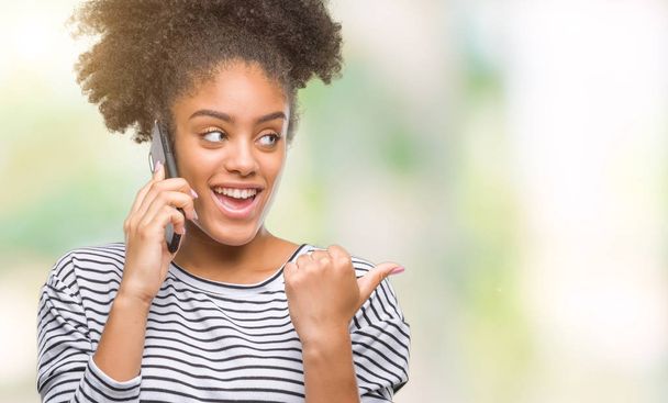 Молодая афро-американка разговаривает по телефону на изолированном фоне указывая и показывая пальцем вверх в сторону со счастливым лицом улыбаясь
 - Фото, изображение