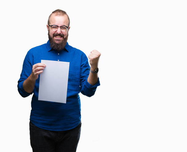 Young hipster man wearing glasses and holding blank paper over isolated background screaming proud and celebrating victory and success very excited, cheering emotion - Photo, Image