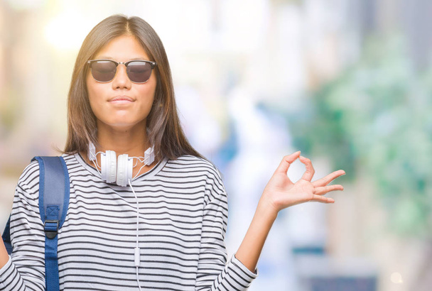 Young asian student woman wearing headphones and backpack over isolated background relax and smiling with eyes closed doing meditation gesture with fingers. Yoga concept. - Foto, imagen
