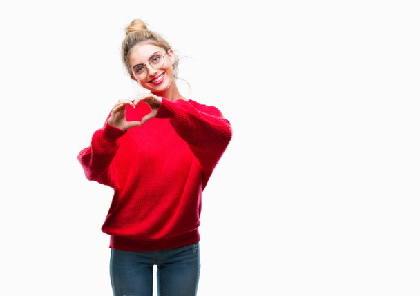 Young beautiful blonde woman wearing red sweater and glasses over isolated background smiling in love showing heart symbol and shape with hands. Romantic concept. - 写真・画像