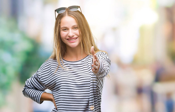 Young beautiful blonde woman wearing sunglasses over isolated background smiling friendly offering handshake as greeting and welcoming. Successful business. - Photo, Image