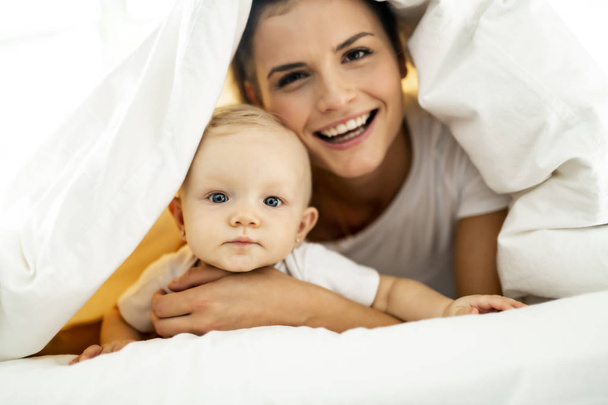 Portrait of young mother with infant baby girl liying on the bed covered with a white blanket - Foto, imagen