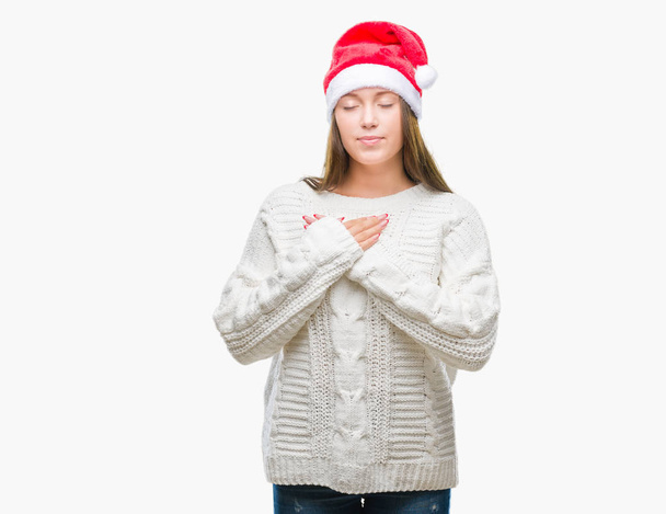 Молодая красивая кавказка в рождественской шляпе на изолированном фоне улыбается руками на груди с закрытыми глазами и благодарным жестом на лице. Концепция здоровья
. - Фото, изображение