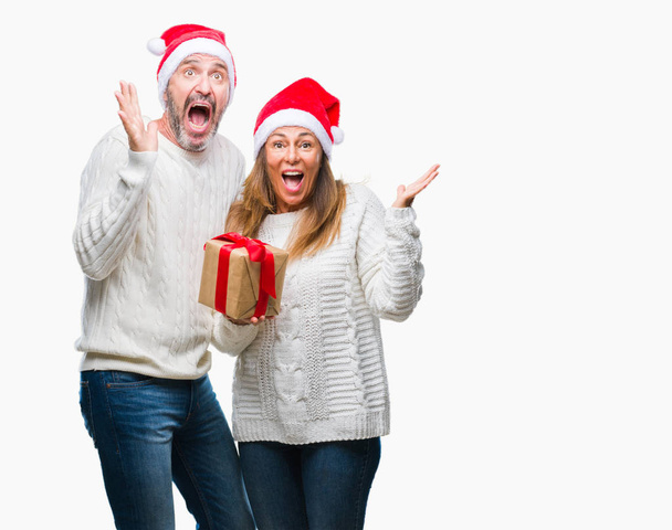 Pareja hispana de mediana edad vistiendo sombrero de navidad y sosteniendo el regalo sobre un fondo aislado muy feliz y emocionado, expresión ganadora celebrando la victoria gritando con una gran sonrisa y levantando las manos
 - Foto, Imagen