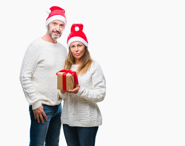 Pareja hispana de mediana edad que usa sombrero de Navidad y sostiene el regalo sobre un fondo aislado con una expresión confiada en el pensamiento serio de la cara inteligente
 - Foto, Imagen
