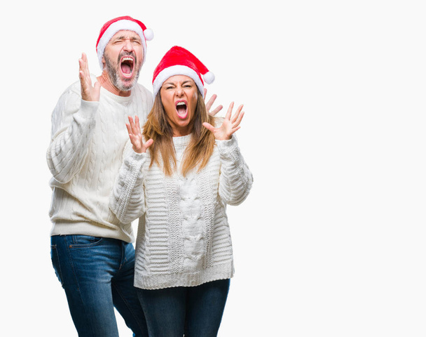 Middle age hispanic couple wearing christmas hat over isolated background crazy and mad shouting and yelling with aggressive expression and arms raised. Frustration concept. - Foto, imagen