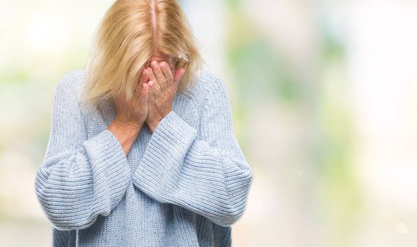 Mujer rubia de mediana edad que usa suéter de invierno sobre un fondo aislado con expresión triste que cubre la cara con las manos mientras llora. Concepto de depresión
. - Foto, imagen