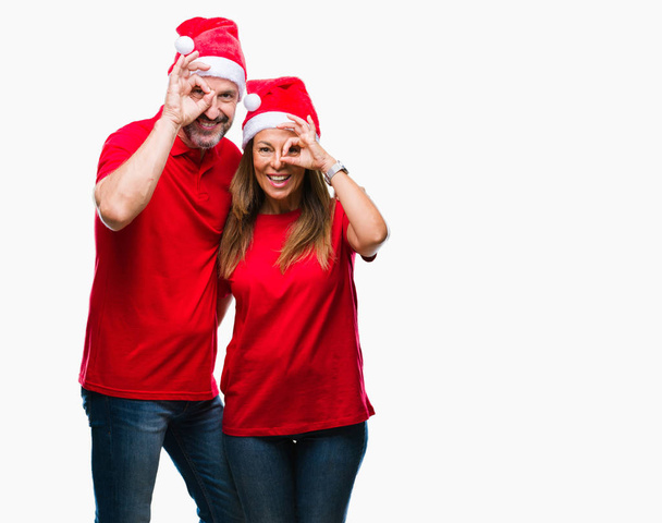 Латиноамериканская пара средних лет в рождественской шляпе на изолированном фоне делает хорошо жест с улыбкой на руке, глаза смотрят сквозь пальцы с счастливым лицом
. - Фото, изображение