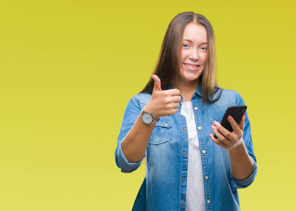 Νεαρή όμορφη γυναίκα Καυκάσιος γραπτών μηνυμάτων που στέλνει το μήνυμα χρησιμοποιώντας το smartphone πέρα από το απομονωμένο υπόβαθρο ευτυχισμένη με μεγάλο χαμόγελο κάνει εντάξει σήμα, τον αντίχειρα επάνω με τα δάχτυλα, καλό σημάδι - Φωτογραφία, εικόνα