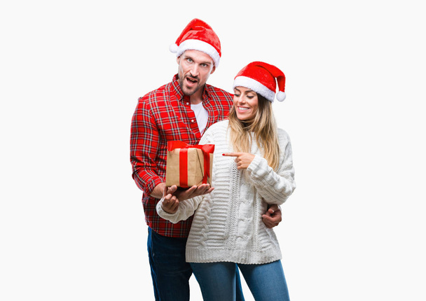 Jong koppel in liefde dragen kerst hoed en bedrijf aanwezig over geïsoleerde achtergrond erg blij met de hand en de vinger te wijzen - Foto, afbeelding