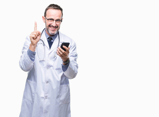 Bliski wieku przy użyciu smartfona przez SMS-y starszy lekarz sędziwy mężczyzna na białym tle zaskoczony z pomysłu lub pytanie palcem wskazującym z szczęśliwy twarz, numer jeden - Zdjęcie, obraz