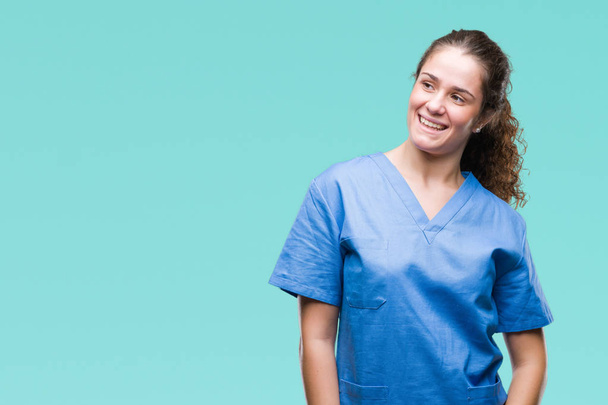 Jeune fille brune médecin portant infirmière ou chirurgien uniforme sur fond isolé regardant de côté avec le sourire sur le visage, expression naturelle. Rire confiant
. - Photo, image