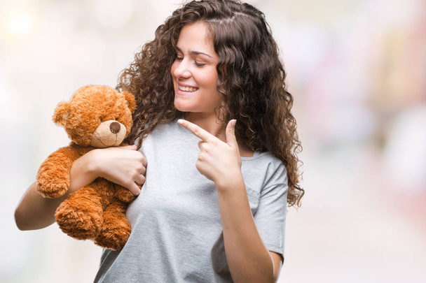 Молода брюнетка дівчина тримає плюшевого ведмедя над ізольованим тлом дуже щаслива, вказуючи рукою і пальцем
 - Фото, зображення