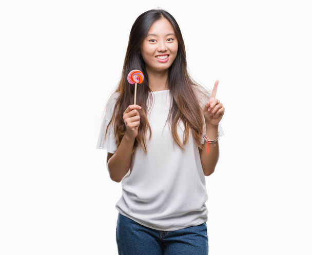 Joven mujer asiática comiendo caramelos de piruleta sobre fondo aislado sorprendido con una idea o pregunta señalando el dedo con la cara feliz, número uno
 - Foto, imagen