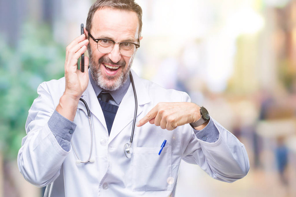 Старший седовласый врач средних лет разговаривает по смартфону на изолированном фоне с удивленным лицом, указывающим пальцем на себя
 - Фото, изображение