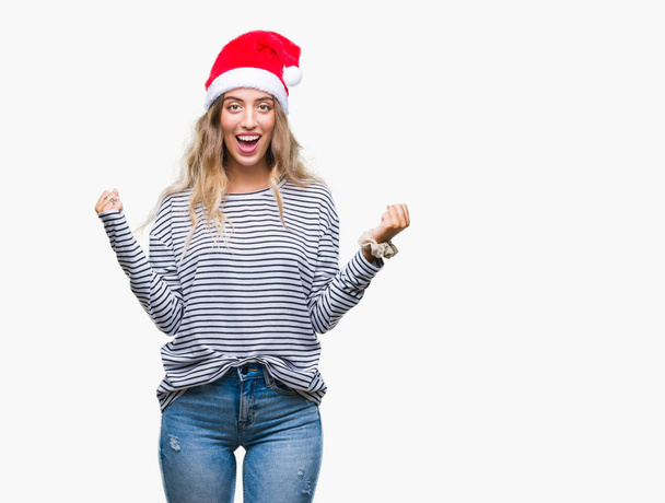 Красивая молодая блондинка в рождественской шляпе на изолированном фоне празднует удивление и удивление от успеха с поднятыми руками и открытыми глазами. Концепция победителя
. - Фото, изображение