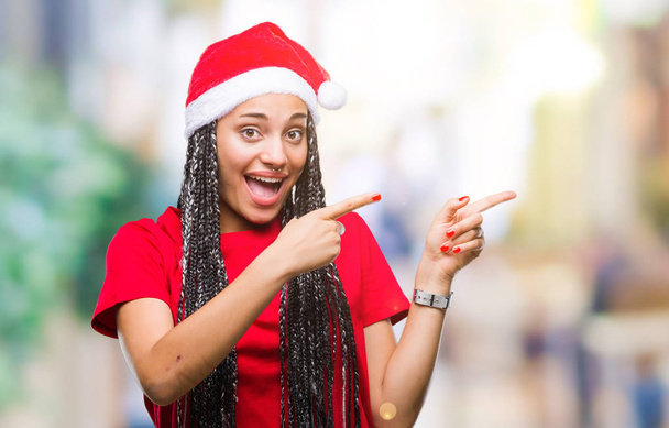 Mladá pletené vlasy africká americká dívka, která nosí vánoční čepice izolované pozadí s úsměvem a při pohledu na fotoaparát s oběma rukama a prsty přejdete na stranu. - Fotografie, Obrázek