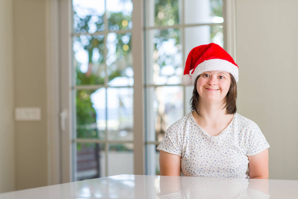Σύνδρομο Down γυναίκα στο σπίτι φορώντας καπέλο Χριστουγέννων με ένα χαρούμενο πρόσωπο στέκονται και να χαμογελούν με αυτοπεποίθηση χαμόγελο δείχνει τα δόντια - Φωτογραφία, εικόνα