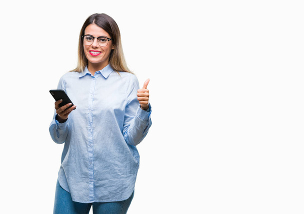 Jonge mooie zakelijke vrouw SMS bericht met smartphone via geïsoleerd achtergrond blij met de grote glimlach doen ok teken, duim omhoog met vingers, uitstekende teken - Foto, afbeelding