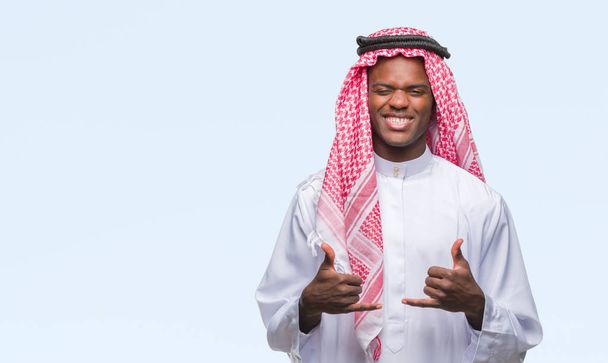 Νεαρός Αραβικά αφρικανικά άνδρας φορώντας παραδοσιακές keffiyeh πάνω από το απομονωμένο ιστορικό σημάδι επιτυχίας κάνει θετική κίνηση με το χέρι, thumbs up χαμογελαστός και χαρούμενος. Κοιτάζοντας την κάμερα με χαρούμενη έκφραση, χειρονομία νικητής. - Φωτογραφία, εικόνα