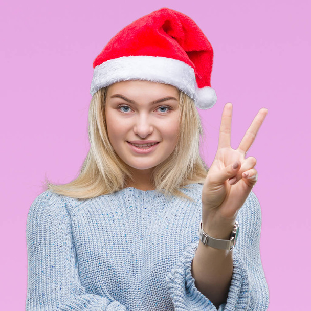 Fiatal kaukázusi nő, fárasztó karácsonyi kalap alatt elszigetelt háttér mosolyogva kacsintott a kamera csinál a győzelem jele, boldog arc. Két szám. - Fotó, kép