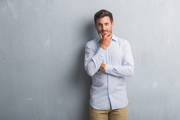 Handsome young business man over grey grunge wall wearing elegant shirt looking confident at the camera with smile with crossed arms and hand raised on chin. Thinking positive. - Photo, image