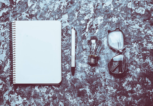 The creative workspace of the writer is inspiring to create. I have an idea. Notepad, pen, incandescent bulb, glasses on a concrete table. Top view. Flat lay. - Photo, Image