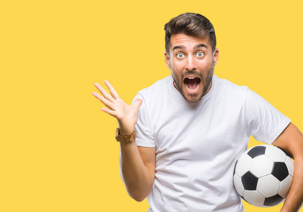 Mladý pohledný muž drží fotbal fotbalový míč nad, samostatný pozadí velmi šťastný a nadšený, vítěz výraz slaví vítězství křičet s velkým úsměvem a pozvedl ruce - Fotografie, Obrázek