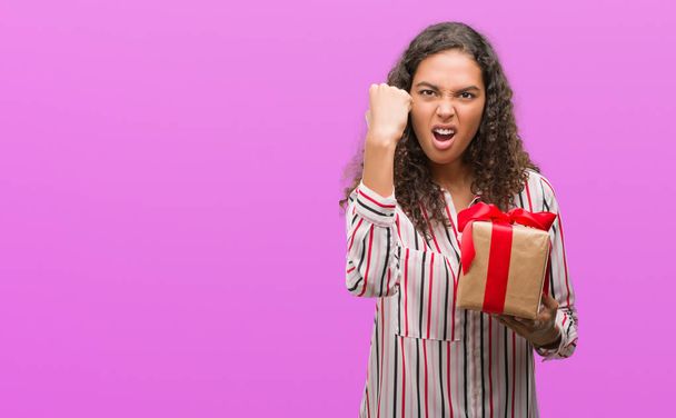 Young hispanic woman in love holding present annoyed and frustrated shouting with anger, crazy and yelling with raised hand, anger concept - Fotoğraf, Görsel