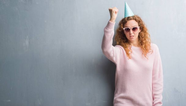 Jonge roodharige vrouw over grijs grunge muur dragen verjaardag cap boos en gefrustreerd schreeuwen met woede, gek en schreeuwen met opgeheven hand, woede concept - Foto, afbeelding
