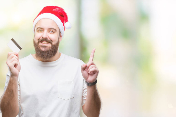 Молодий білий чоловік у різдвяному капелюсі тримає кредитну картку над ізольованим тлом здивований ідеєю або питанням, що вказує пальцем щасливе обличчя, номер один
 - Фото, зображення