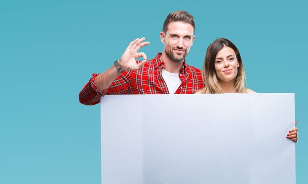 Young couple together holding blank banner over isolated background doing ok sign with fingers, excellent symbol - Valokuva, kuva