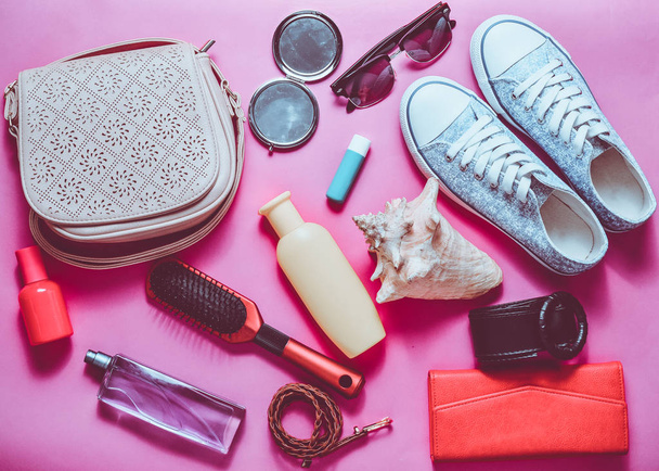 Wat is in de women's zak? Gaande van een reis. Girly modieuze lente en zomer accessoires: sneakers, cosmetica, schoonheid en hygiëne producten, een zak, zonnebril op een roze pastel achtergrond. Bovenaanzicht. - Foto, afbeelding
