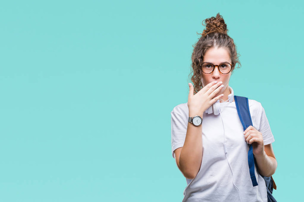 Young brunette student girl wearing backpack and headphones over isolated background shocked covering mouth with hands for mistake. Secret concept. - Photo, Image