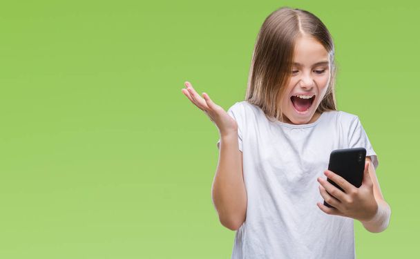 junges schönes Mädchen sendet SMS per Smarpthone über isolierten Hintergrund sehr glücklich und aufgeregt, Siegerausdruck feiert Sieg schreiend mit breitem Lächeln und erhobenen Händen - Foto, Bild
