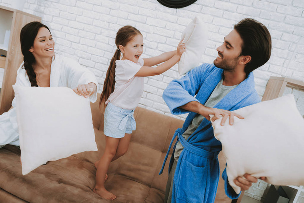 Happy Family Morning. Fight with Pillows Parents and Daughter. Family Resting Together. Morning Fun. Man and Woman in Coats. Smiling Family Resting. Girl Jumping on Sofa. Happy Child and Parents. - Photo, image