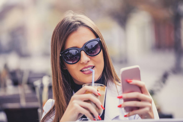 Ücretsiz Wi-Fi zevk. Akıllı tutan neşeli genç kadın telefon sıcak çikolata içme ve Sokak kafede otururken gülümseme ile bakmak - Fotoğraf, Görsel